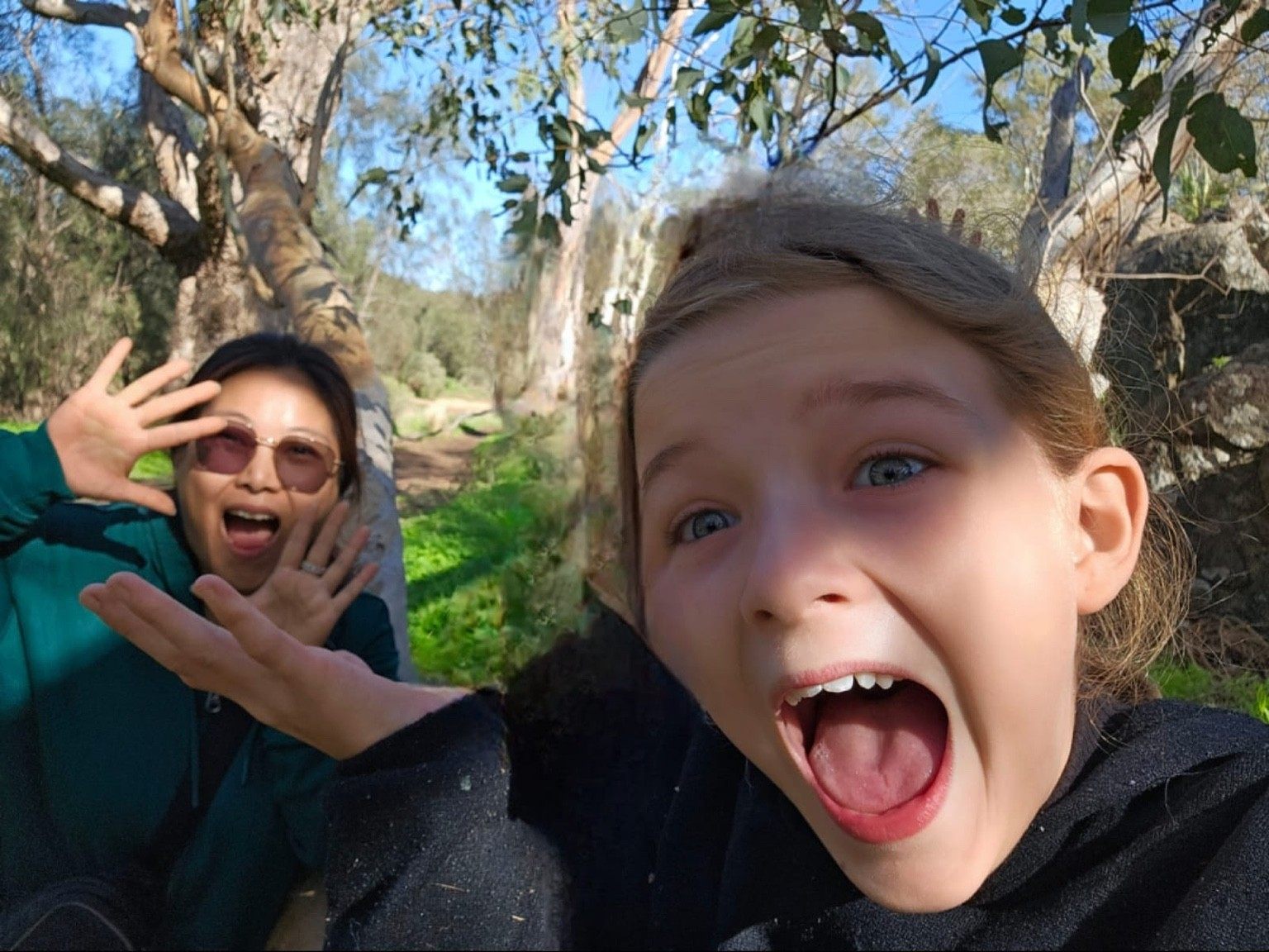 A young girl pulls a silly face for a selfie while a support worker waves and laughs behind her on a sunny bush trail